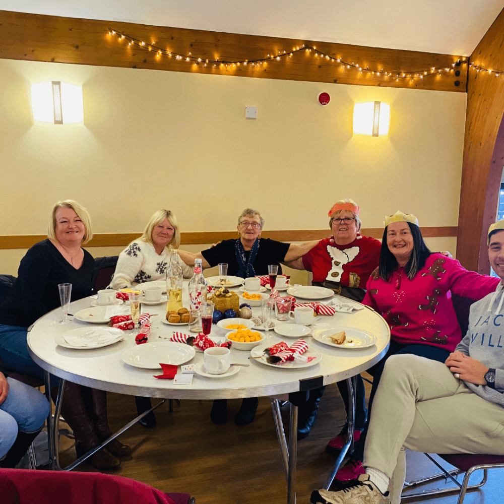 A group of six people seated around a table with drinks and snacks, smiling in a festively decorated room. - Home Instead
