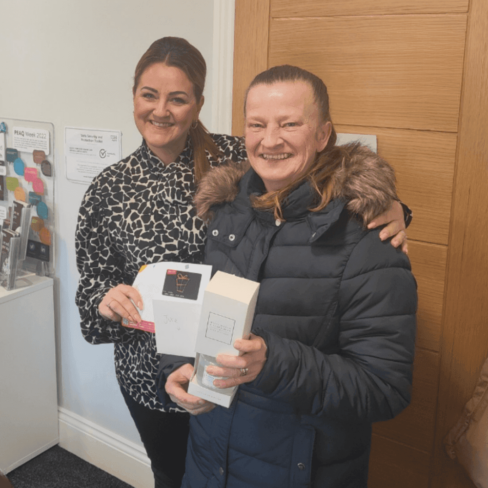 Two women smiling, one holding a small box and card, standing indoors in a casual setting. - Home Instead