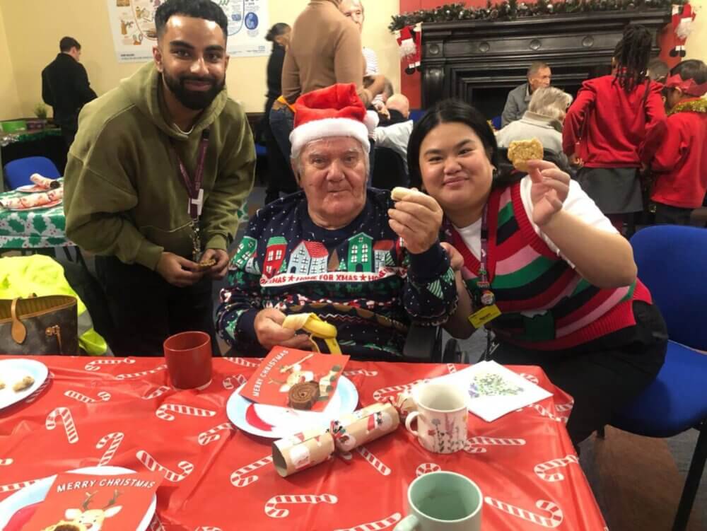 Three people smiling at a festive table, one in a Santa hat and Christmas sweater, displaying holiday treats. - Home Instead