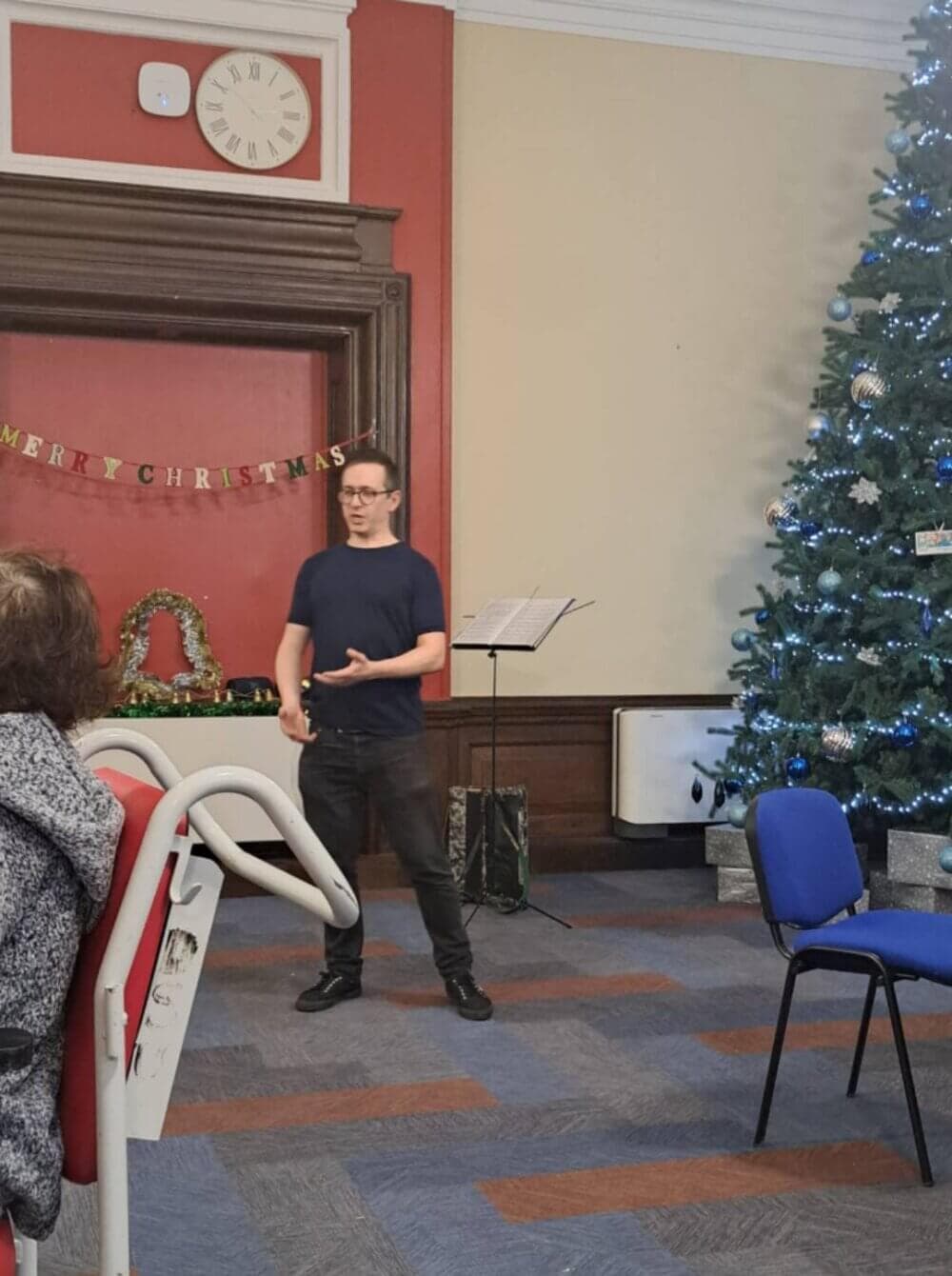 A person speaking in front of a Christmas tree and "Merry Christmas" banner in a decorated room. - Home Instead