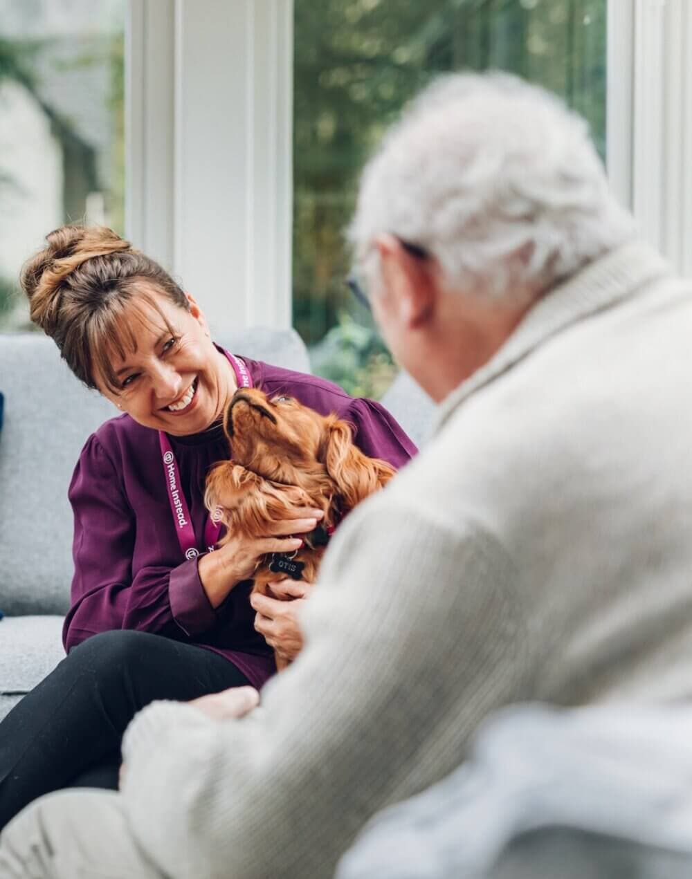 Woman smiling at a man while holding a dog; both sitting in a bright, cozy room. - Home Instead
