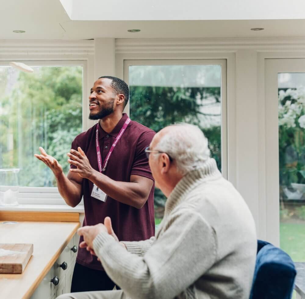 A caregiver in a maroon shirt, smiling and chatting with an elderly man wearing a gray sweater in a bright kitchen. - Home Instead
