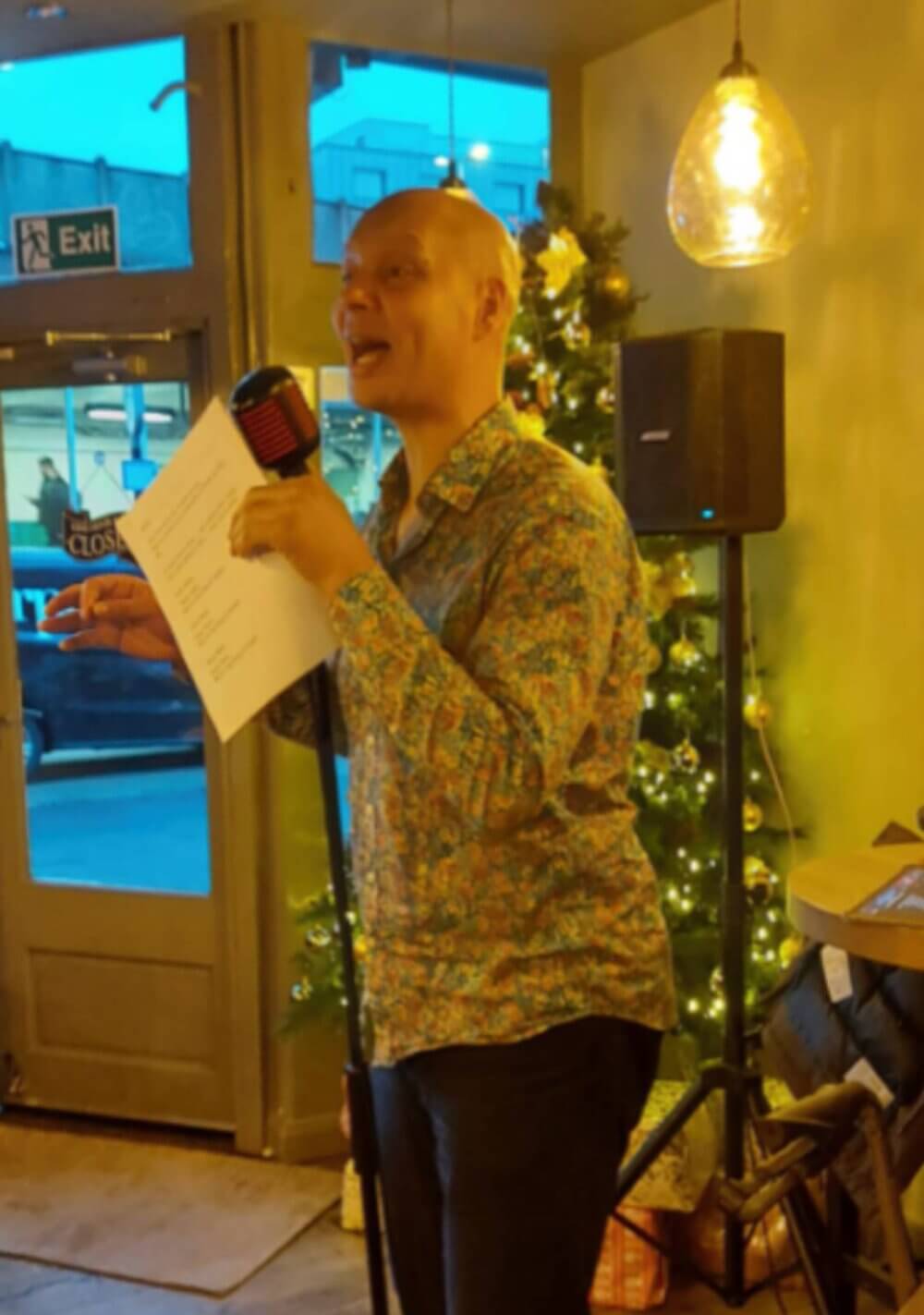 Person speaking into a microphone while holding papers in a cozy room with a lit Christmas tree and a large window in the background. - Home Instead