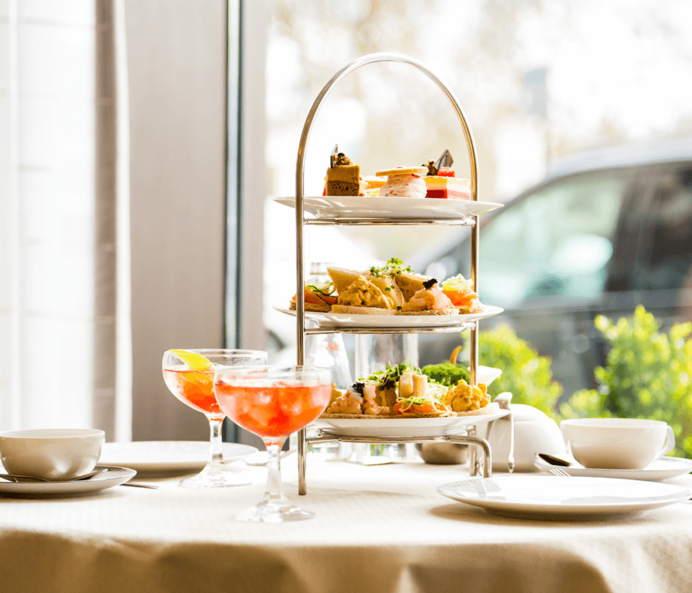 A three-tiered serving stand with assorted pastries and sandwiches, alongside two cocktails, on a table by a window. - Home Instead