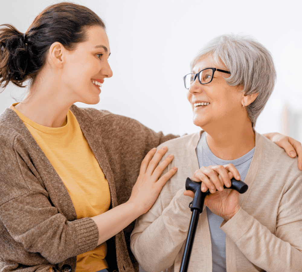Young woman smiling and touching the shoulder of an elderly woman with glasses and a cane, both looking at each other warmly. - Home Instead