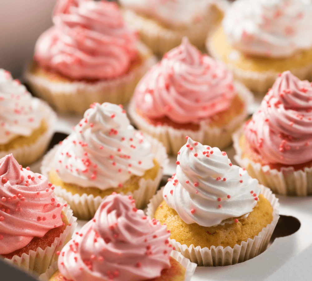 Close-up of cupcakes topped with pink and white swirled frosting and red sprinkles arranged in a box. - Home Instead