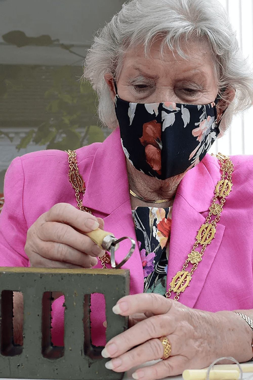 Elderly woman in a floral mask and pink blazer works on a green foam block, wearing a gold chain necklace. - Home Instead
