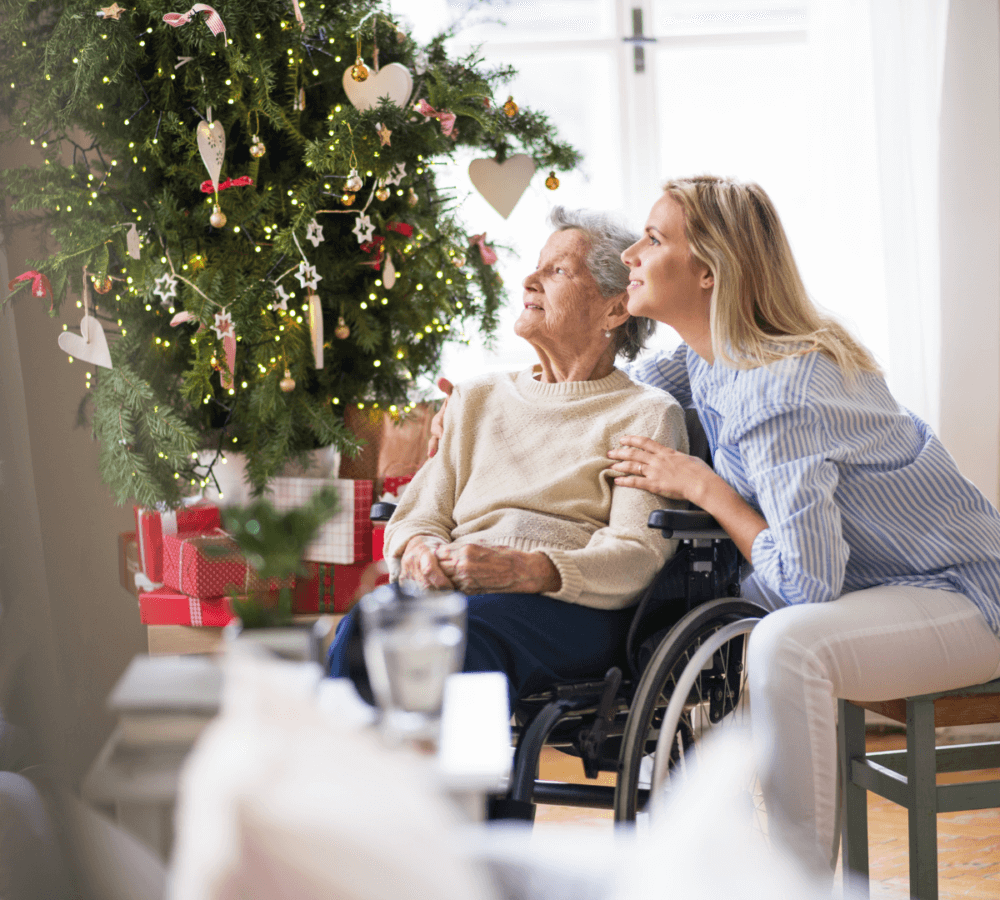 Two women, one elderly in a wheelchair and one younger, look at a decorated Christmas tree with gifts underneath. - Home Instead