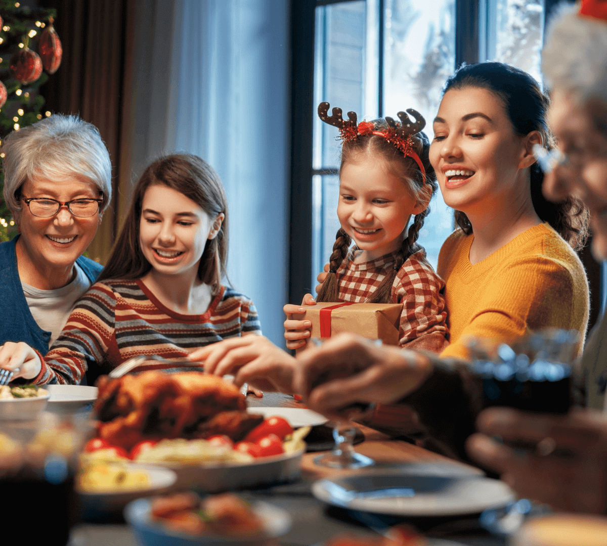 Family gathered around a festive dinner table, smiling and enjoying a holiday meal. A child wears reindeer antlers. - Home Instead