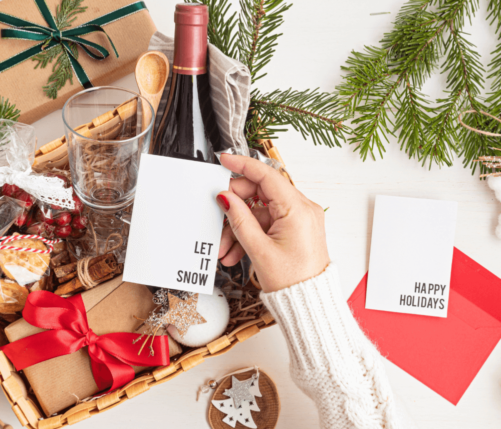 A hand holds a "Let It Snow" card above a festive gift basket. Another card nearby reads "Happy Holidays. - Home Instead
