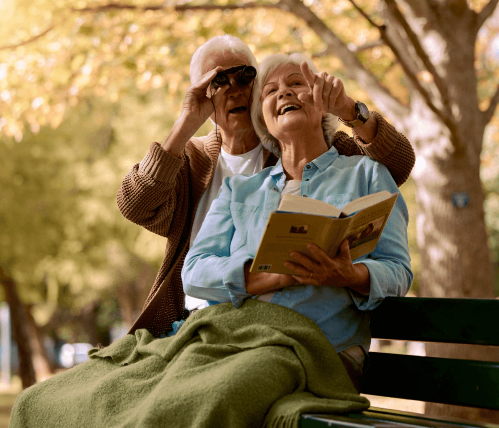 An elderly couple on a bench, the woman reading a book and the man using binoculars, both smiling and pointing ahead. - Home Instead