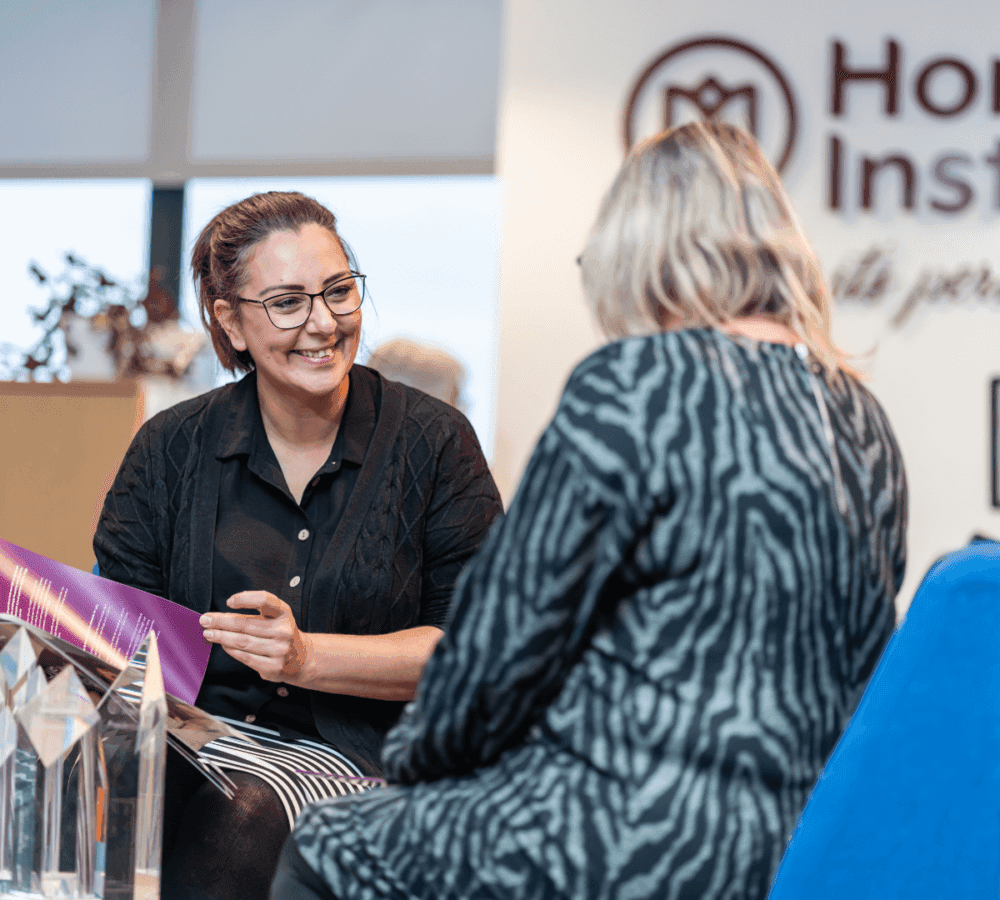 Two women seated and having a conversation, with one showing a folder to the other, in a modern office setting. - Home Instead