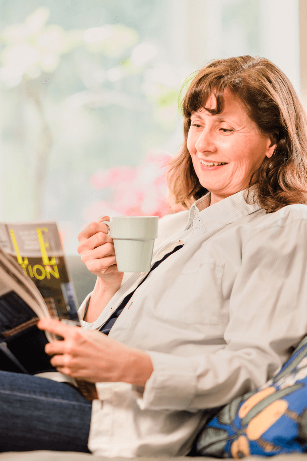 A woman, smiling and sitting on a couch, reads a magazine while holding a coffee mug in a bright, cozy room. - Home Instead