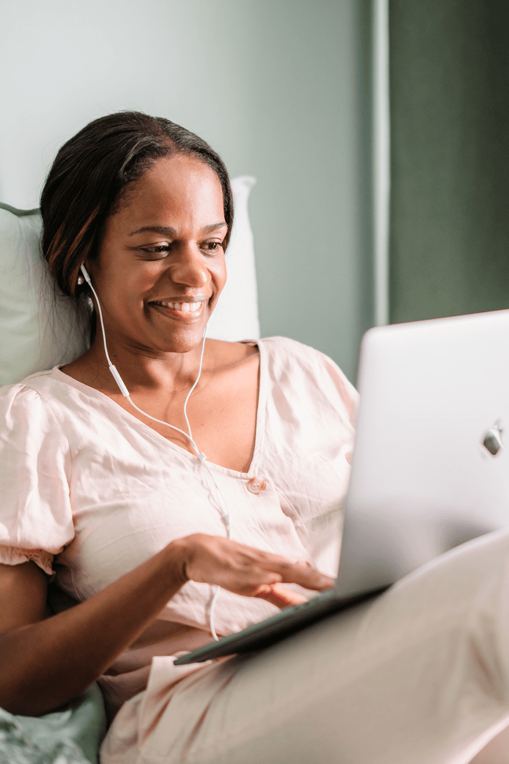 A woman smiles while using a laptop and wearing earphones, sitting comfortably in a casual outfit. - Home Instead