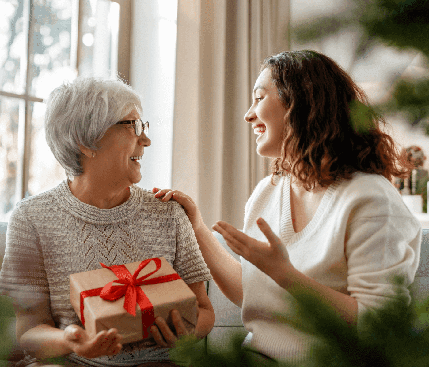Elderly woman and young woman smiling while exchanging a gift wrapped in brown paper with a red ribbon. - Home Instead
