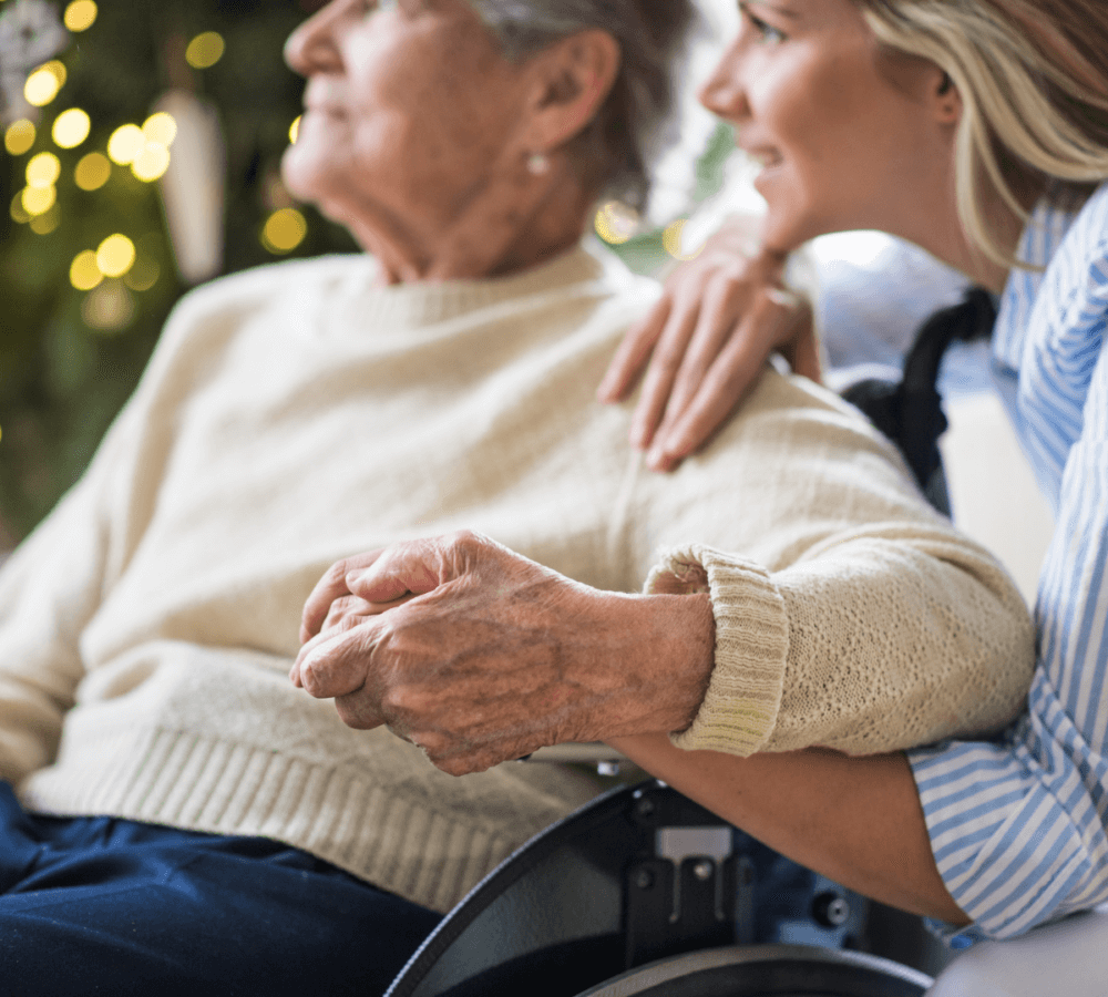 An elderly person in a wheelchair holds hands with a younger person, both smiling and looking off in the same direction. - Home Instead