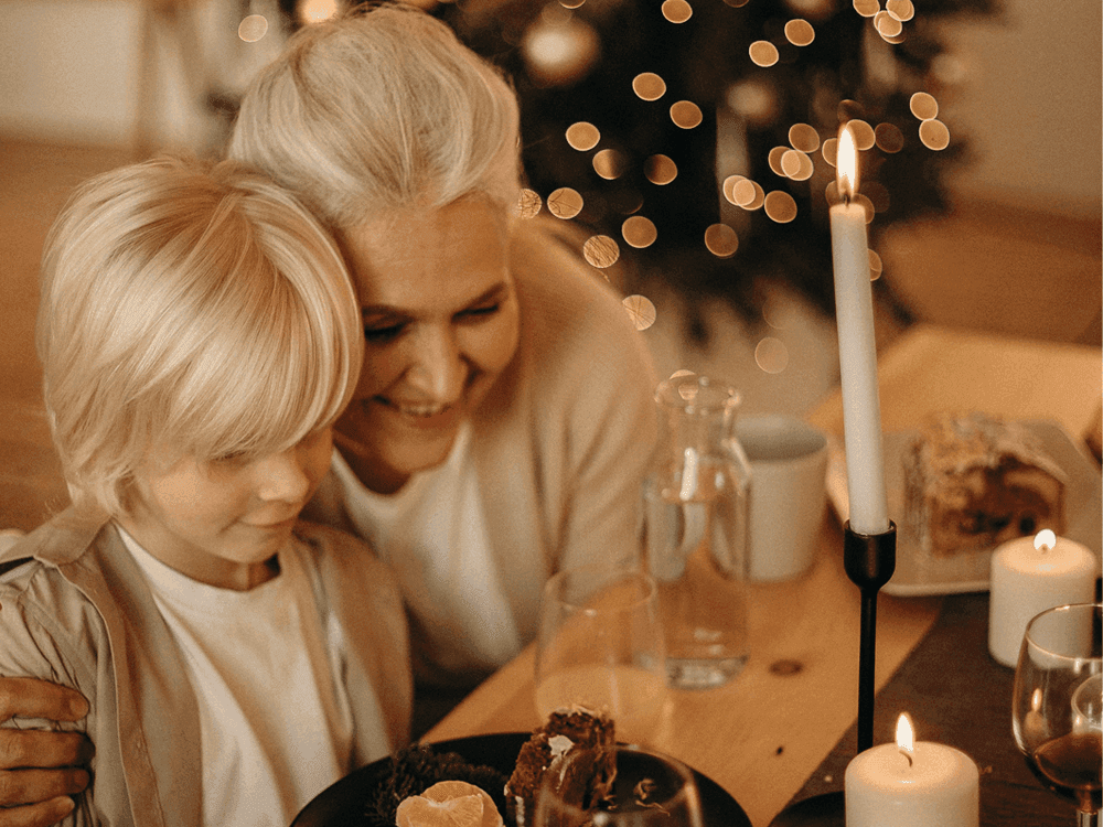 A grandmother and grandchild share a warm embrace at a festive dinner table with candles and holiday lights. - Home Instead