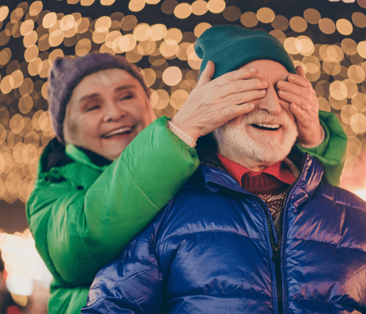 An elderly woman covers an elderly man's eyes playfully, both smiling, under festive string lights. - Home Instead
