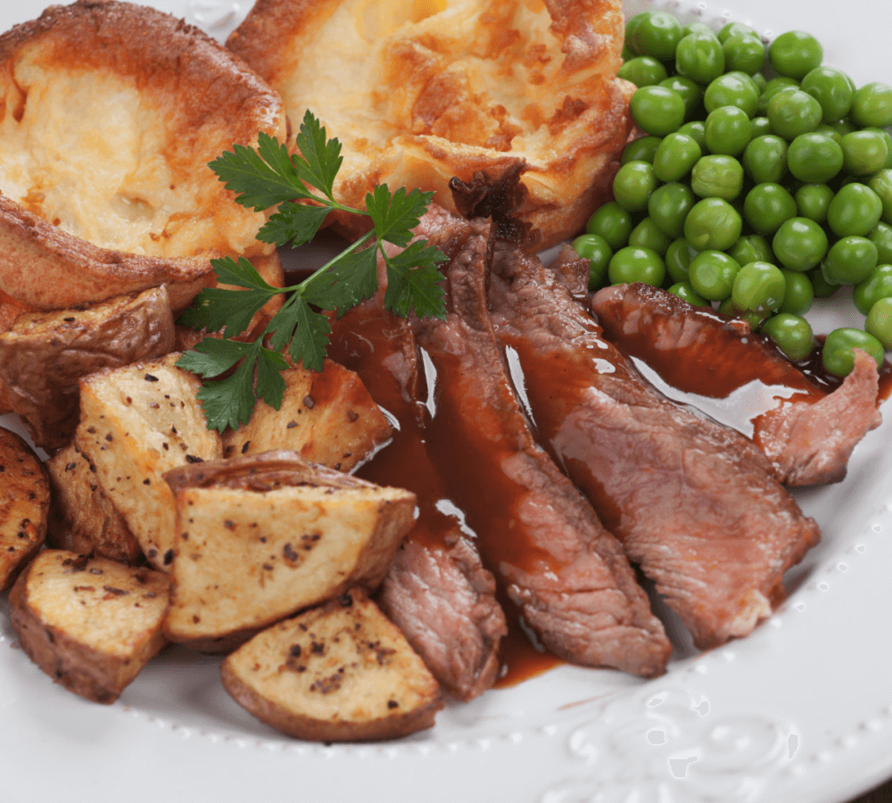 Plate with roasted beef slices, gravy, roast potatoes, Yorkshire puddings, and green peas garnished with parsley. - Home Instead
