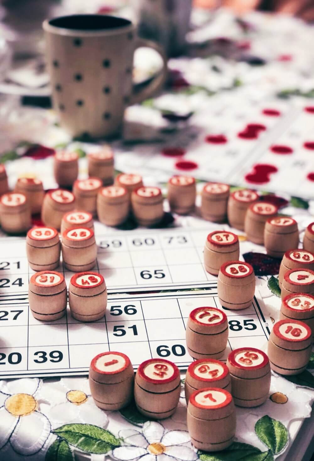 Bingo game in progress with wooden numbered pieces, cards, and a polka dot mug on a floral tablecloth. - Home Instead