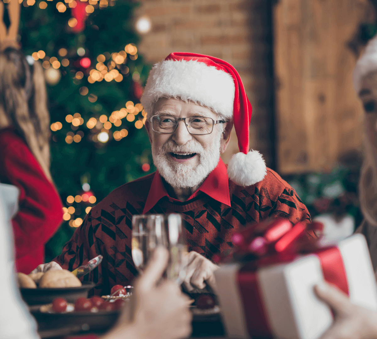 Elderly man wearing Santa hat smiles while dining with family, surrounded by festive decorations and a Christmas tree. - Home Instead