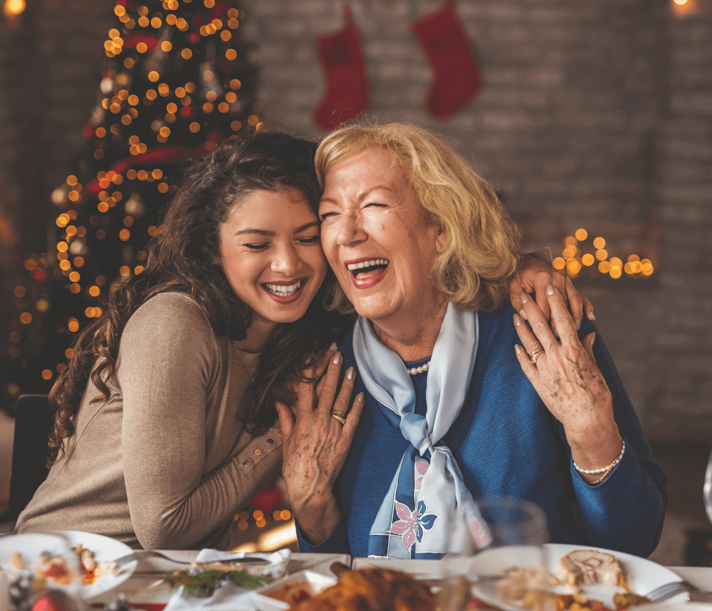 Two women laughing together at a festive table, with a decorated Christmas tree and stockings in the background. - Home Instead