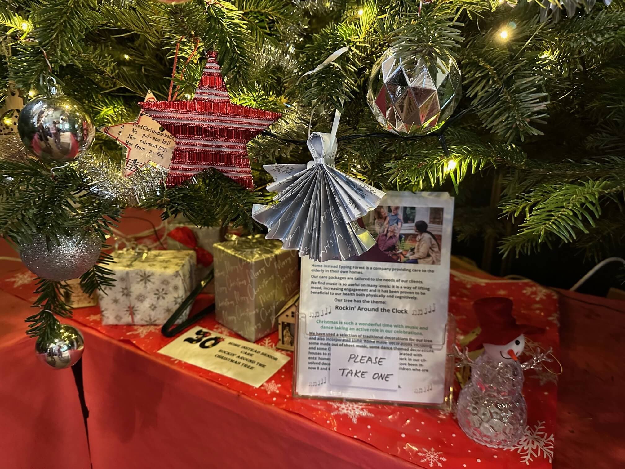 A decorated Christmas tree with ornaments, gifts, and a sign saying "Please take one" on a red cloth-covered table. - Home Instead