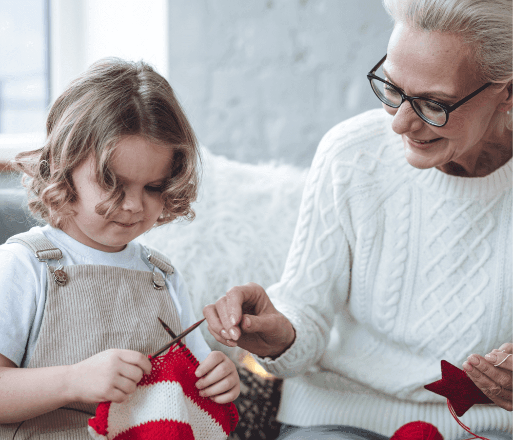 Older woman teaching a child to knit a red and white striped piece, sitting together in a cozy indoor setting. - Home Instead