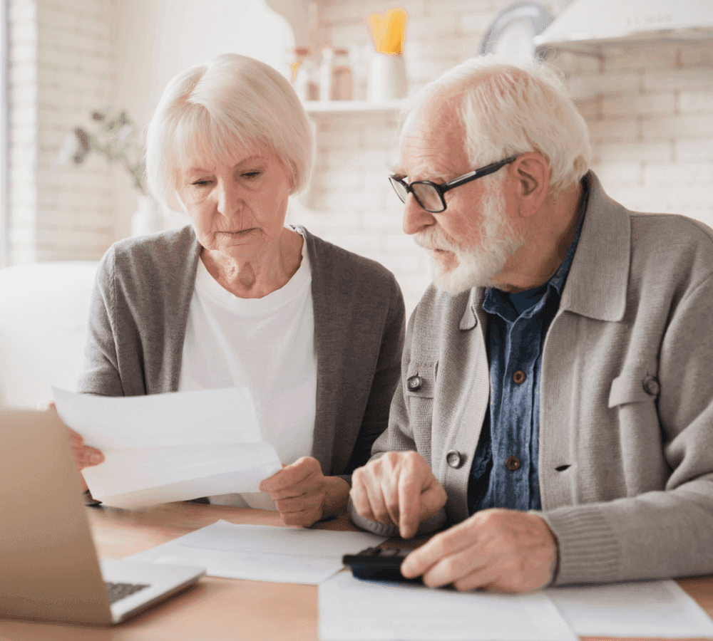 Elderly couple reviewing documents and using a calculator while sitting at a table with a laptop in a brightly lit room. - Home Instead