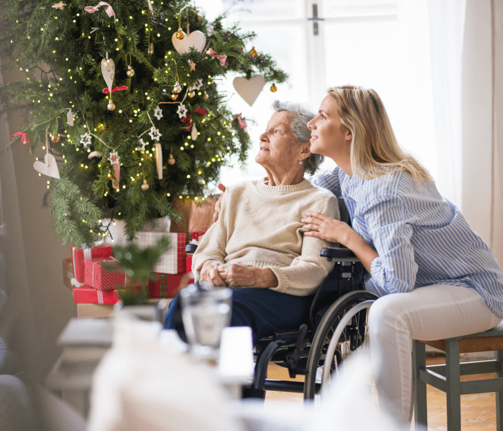 Elderly woman in a wheelchair with a young woman by a decorated Christmas tree, both looking upwards and smiling. - Home Instead