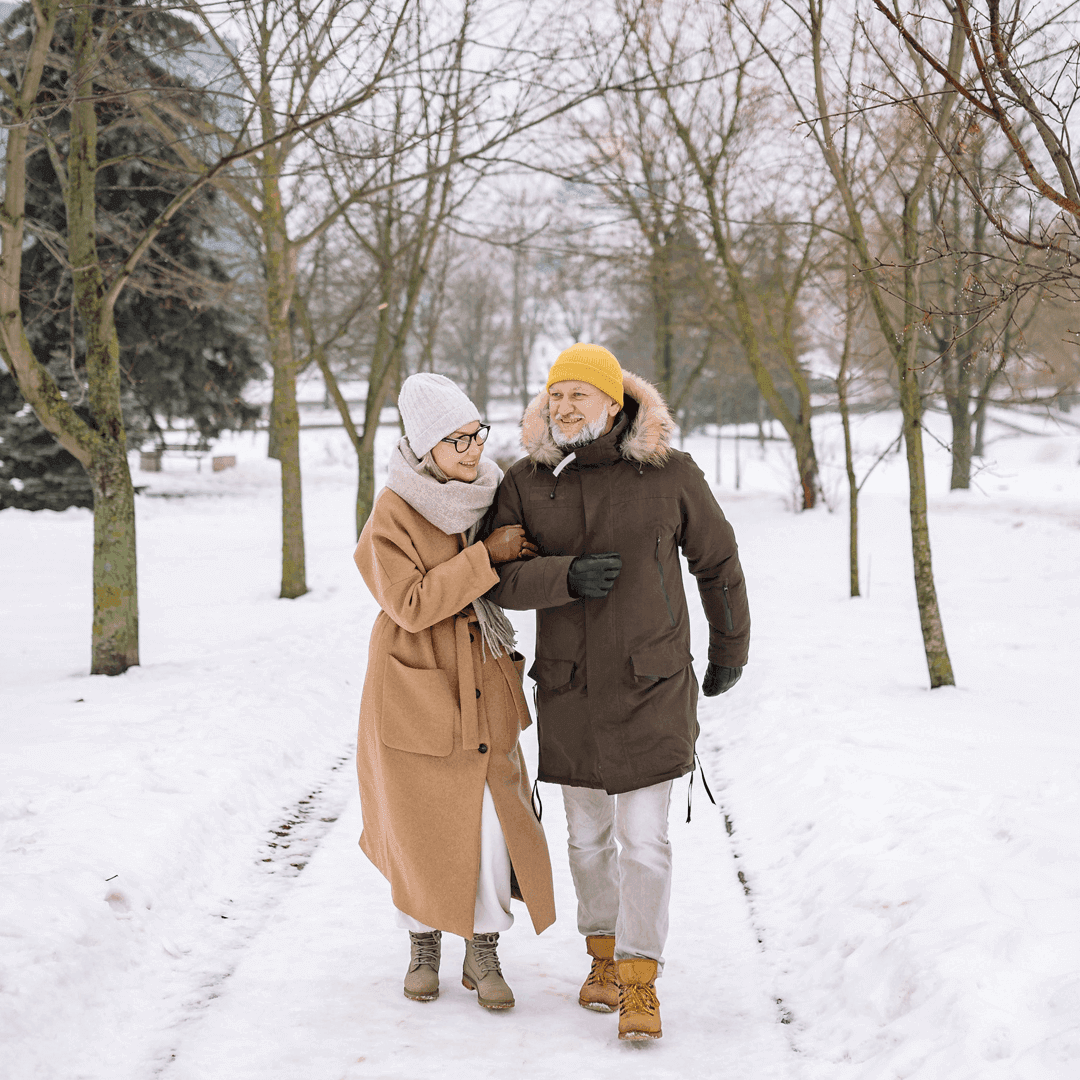 Elderly couple walking through a snowy park, dressed warmly in coats, hats, and scarves, smiling and holding hands. - Home Instead