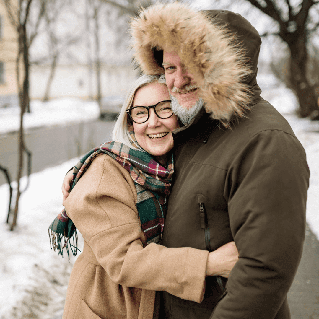 A smiling couple embraces outdoors in winter clothing, with snow on the ground and trees in the background. - Home Instead