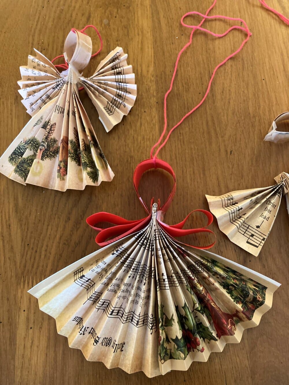 Handmade paper angels crafted from folded music sheets and holiday-themed paper, tied with red strings on a wooden surface. - Home Instead
