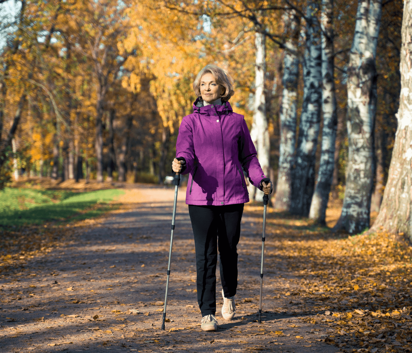 A woman in a purple jacket walks on a leaf-strewn path in a forest, using walking poles, with trees in the background. - Home Instead
