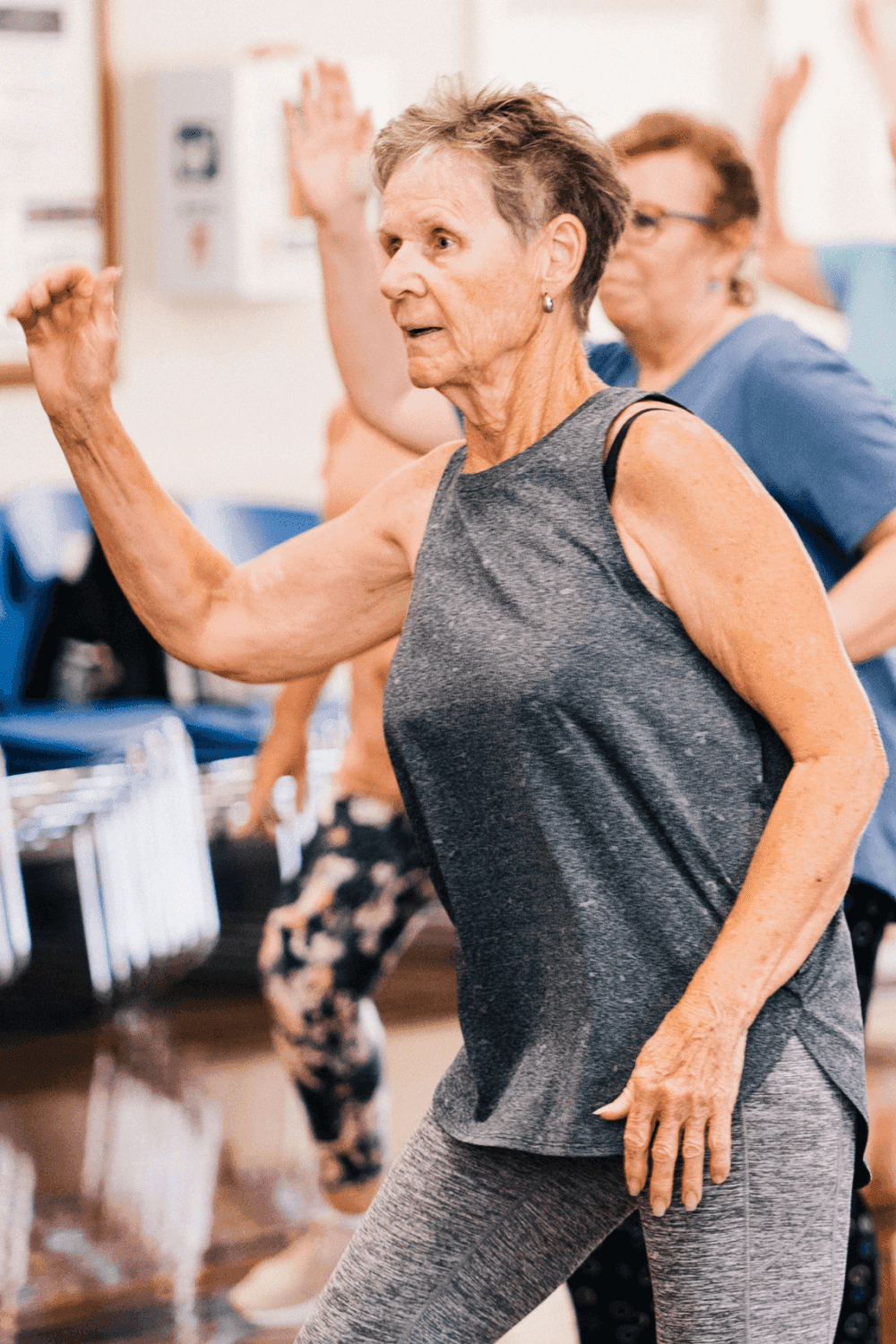 Older adults participating in a group exercise class, focusing on a woman in a gray sleeveless shirt doing an arm movement. - Home Instead