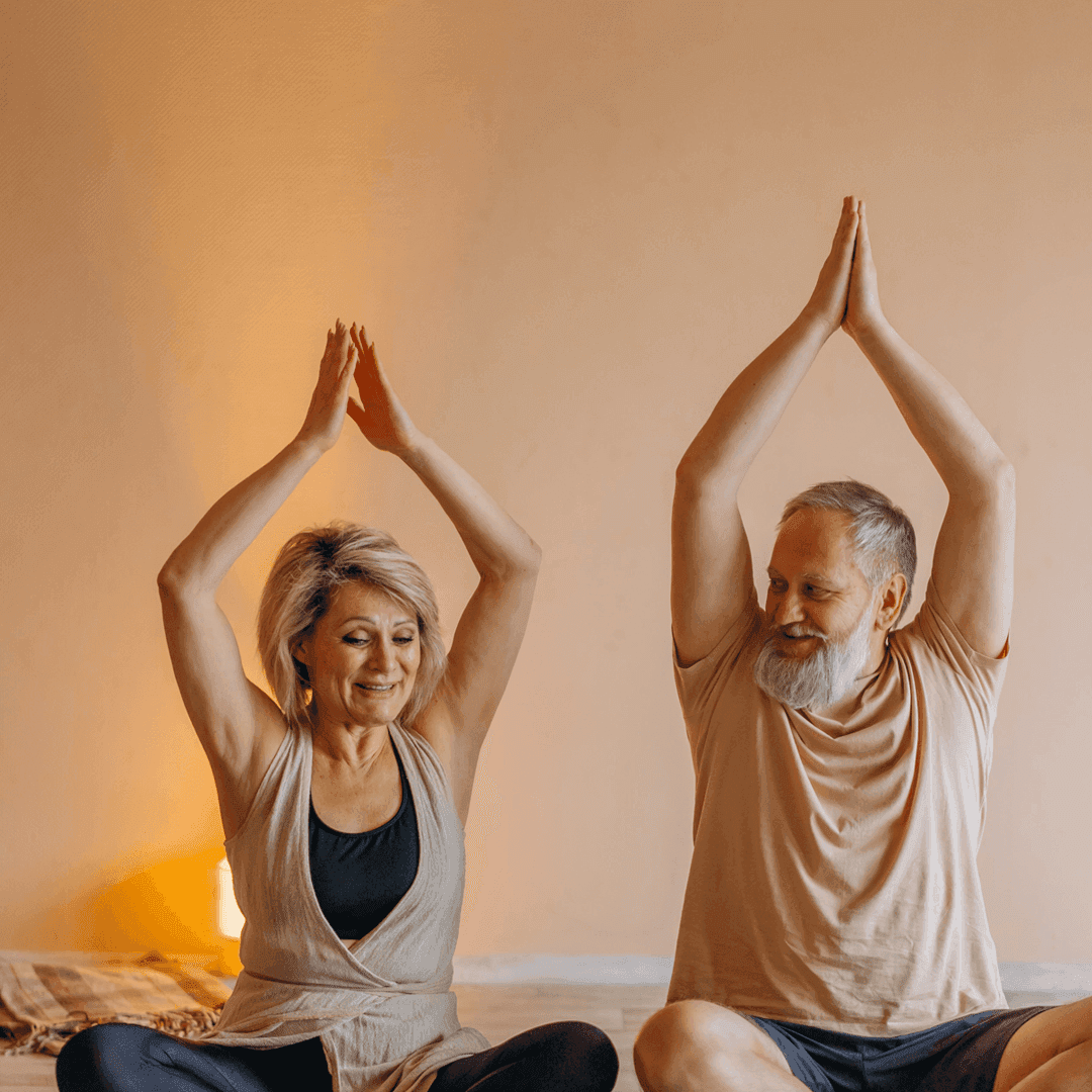 Senior man and woman sitting cross-legged, raising arms over their heads while doing yoga indoors. - Home Instead