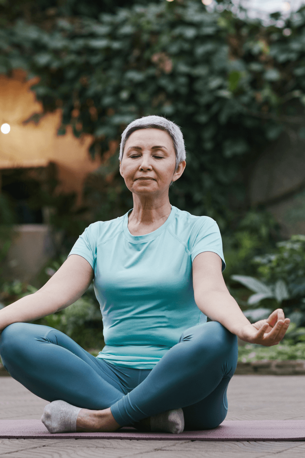 A woman with short gray hair meditates in a seated yoga pose outdoors, with greenery in the background. - Home Instead