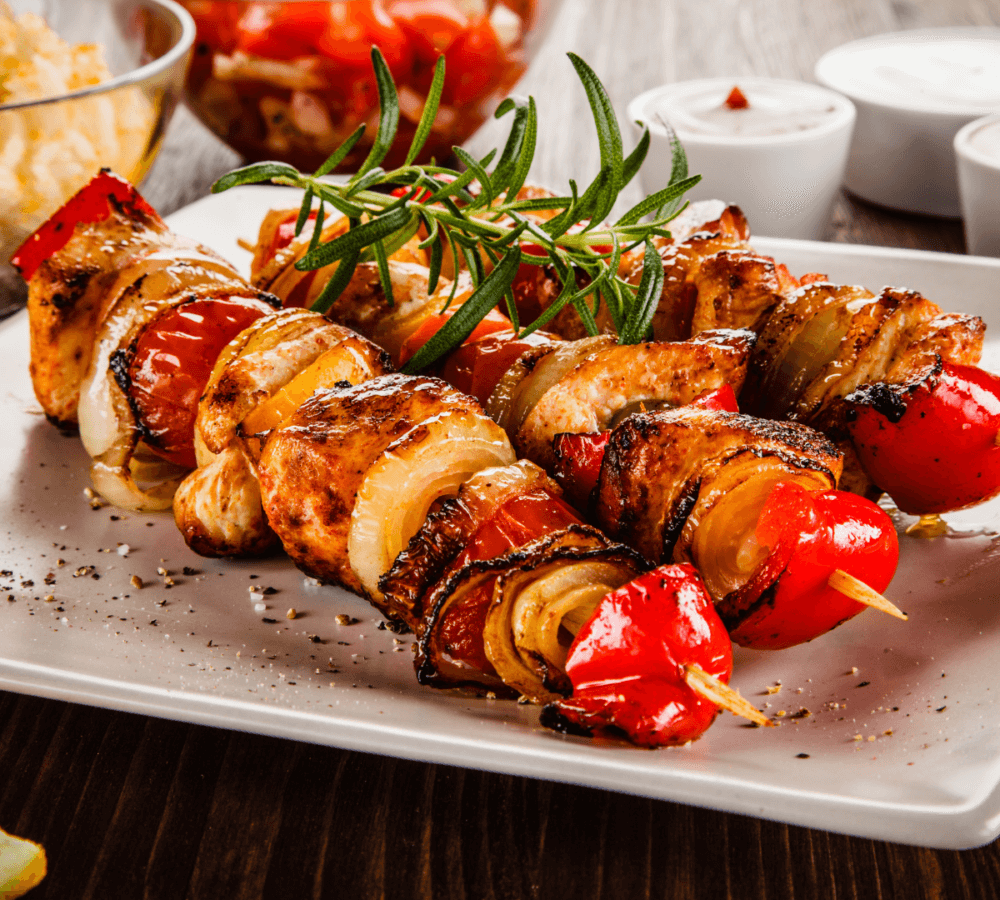 Three grilled chicken and vegetable skewers garnished with rosemary on a white plate, with dips in the background. - Home Instead