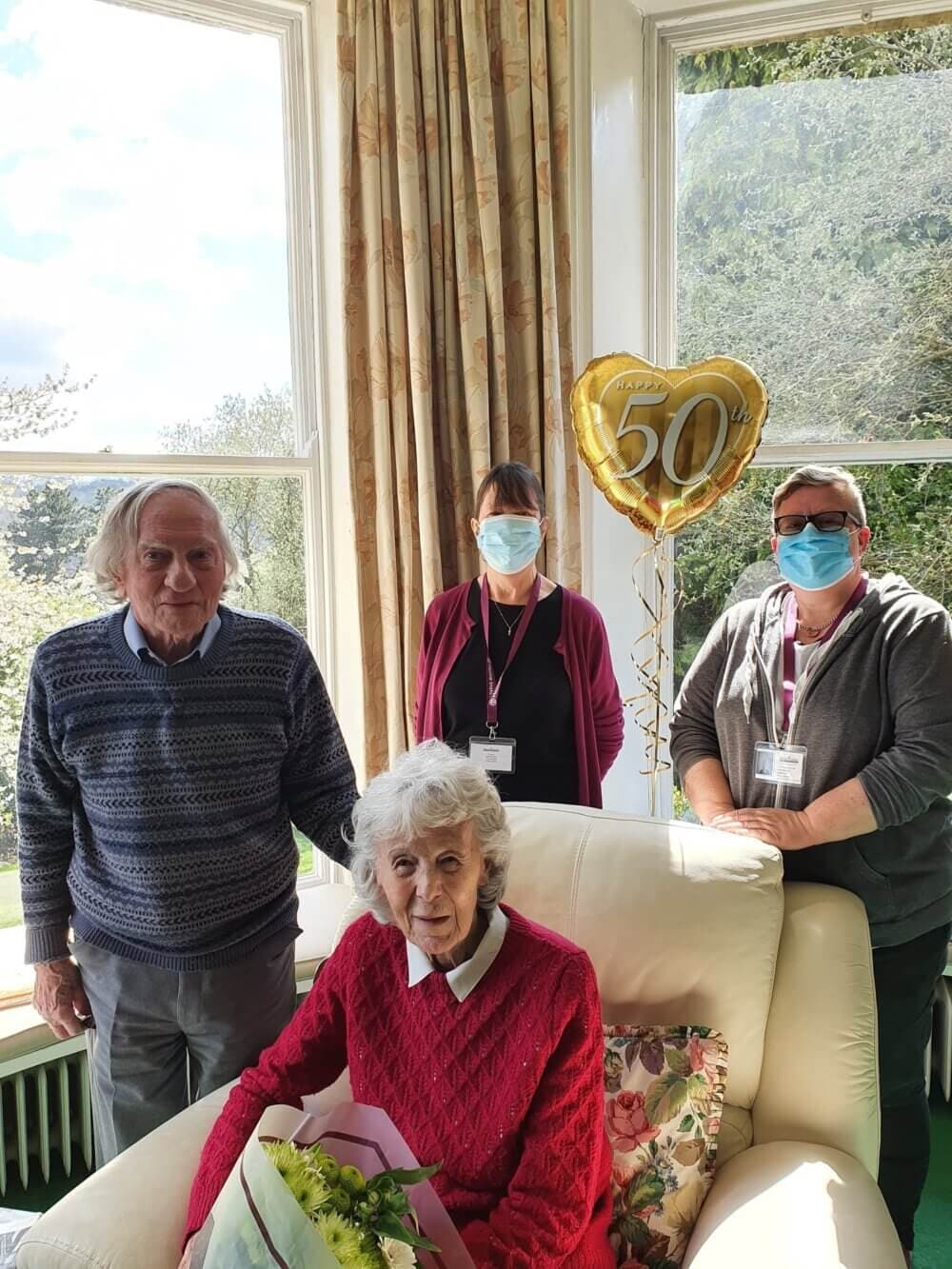 Elderly woman in red sweater with bouquet, seated, flanked by three masked adults, and a 50th-anniversary balloon behind her. - Home Instead
