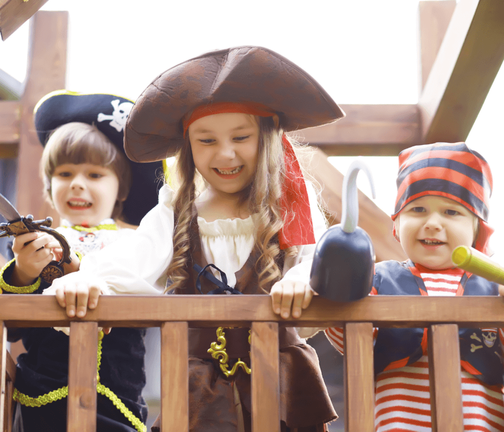 Three young children dressed as pirates smile and play on a wooden play structure. - Home Instead