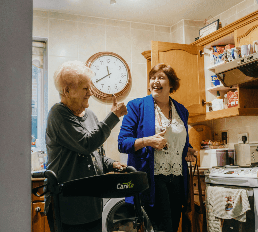 Two women in a kitchen share a laugh, one using a walker. A clock on the wall shows it's past 3 o'clock. - Home Instead