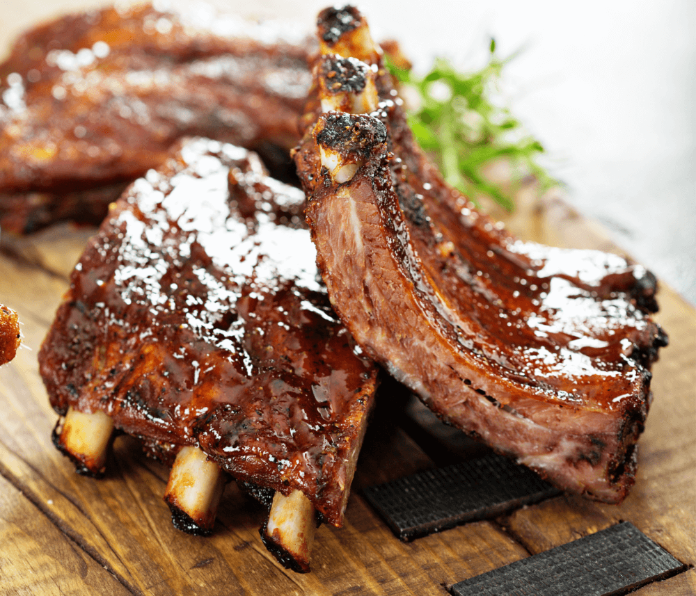 Close-up of glazed barbecue ribs garnished with herbs, served on a wooden board. - Home Instead