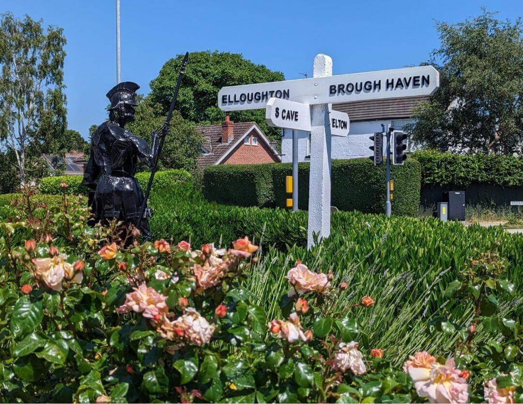 A white signpost in a garden points to Elloughton and Brough Haven; a statue of a knight stands nearby. - Home Instead