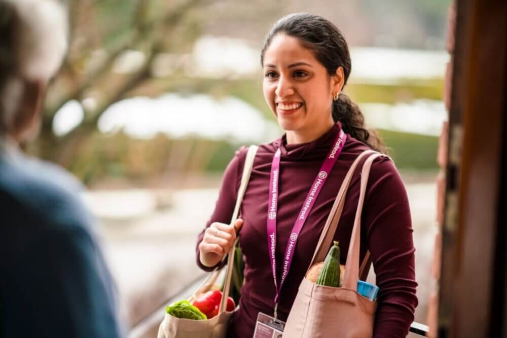 Woman smiling at an older person while carrying grocery bags with vegetables and wearing a name badge. - Home Instead