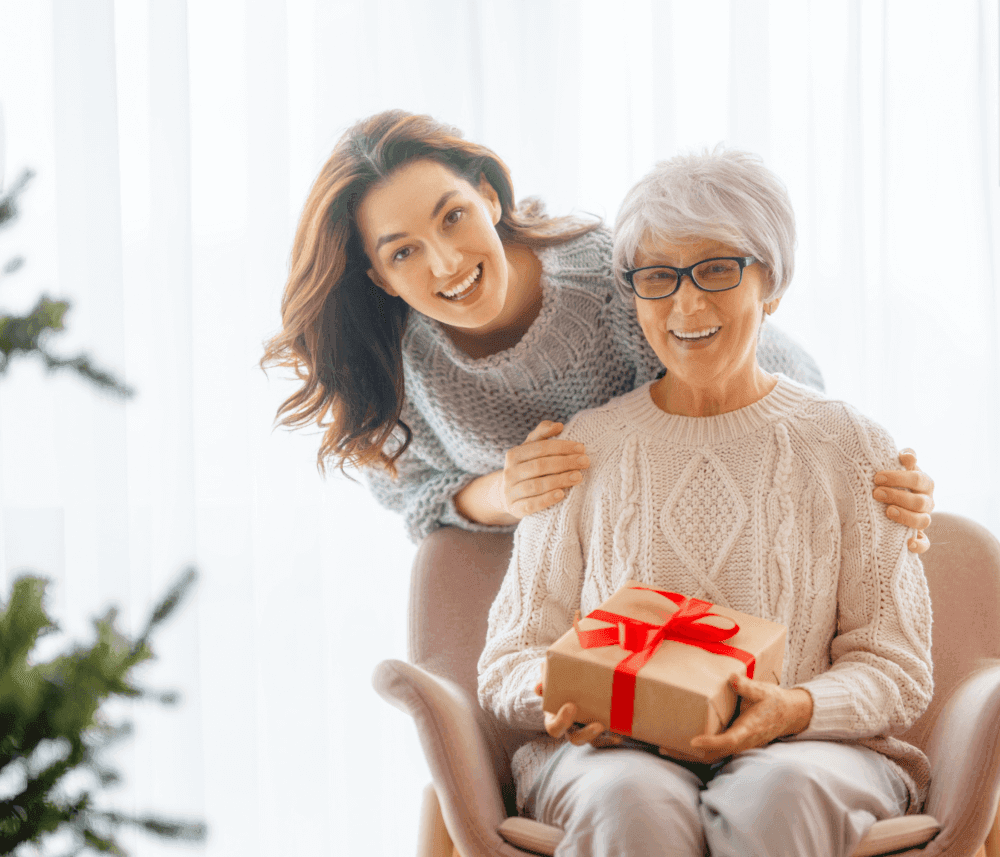 Two women smiling, one seated holding a gift with a red ribbon, the other standing behind her with her hands on her shoulders. - Home Instead