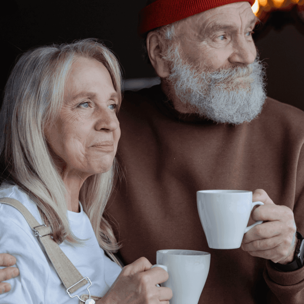 An elderly couple, each holding a cup of coffee, stand closely together, gazing into the distance. - Home Instead