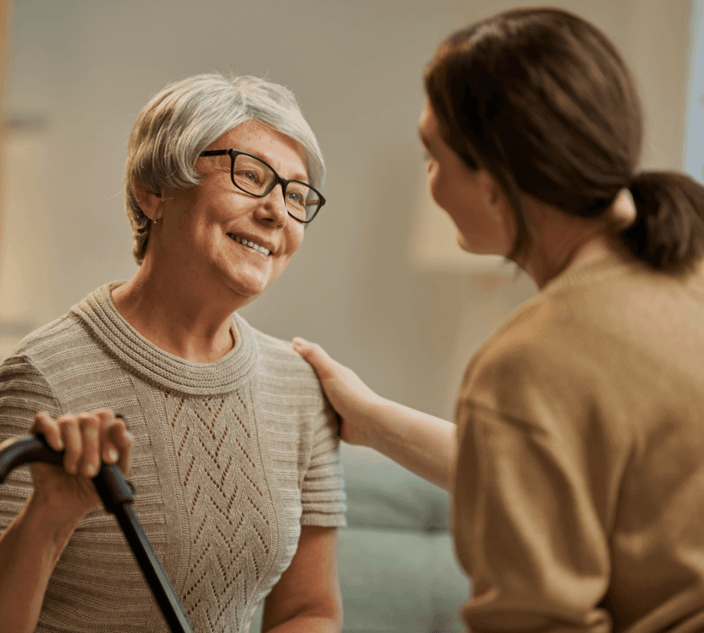 Elderly woman with glasses and a cane smiles at a young woman who is touching her shoulder in a supportive gesture. - Home Instead