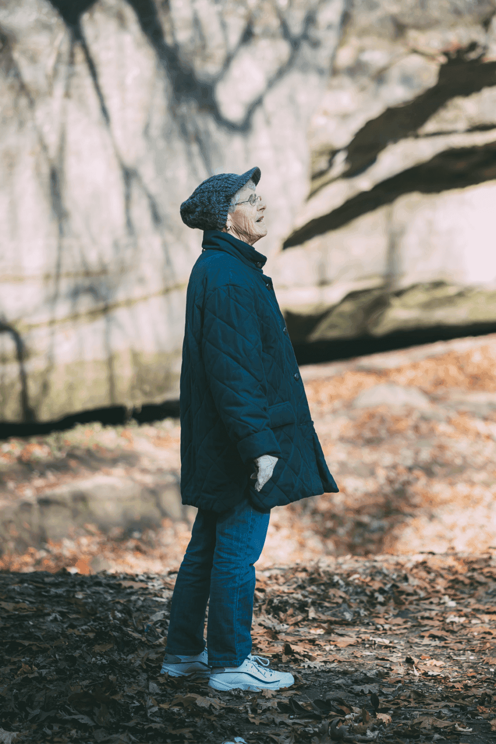 An elderly person in a quilted coat and hat stands in a wooded area looking up, surrounded by fallen leaves. - Home Instead