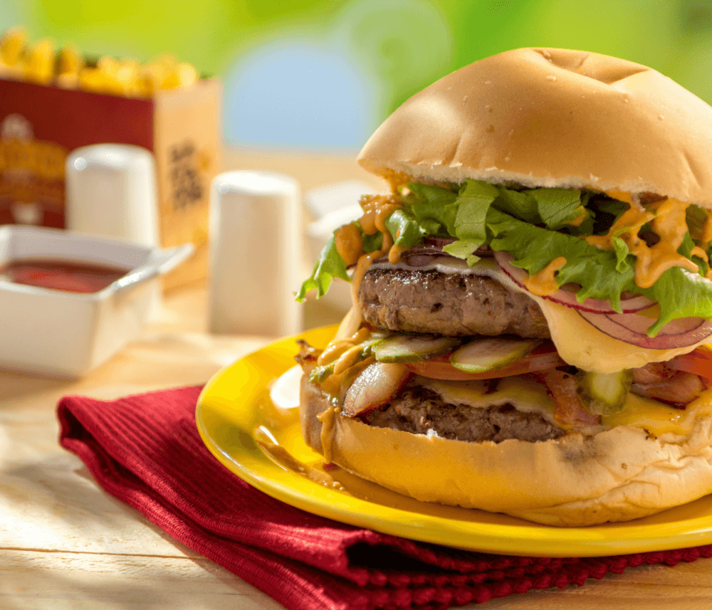 A cheeseburger with lettuce, tomato, pickles, onions, and sauce on a yellow plate next to a red napkin and condiments. - Home Instead