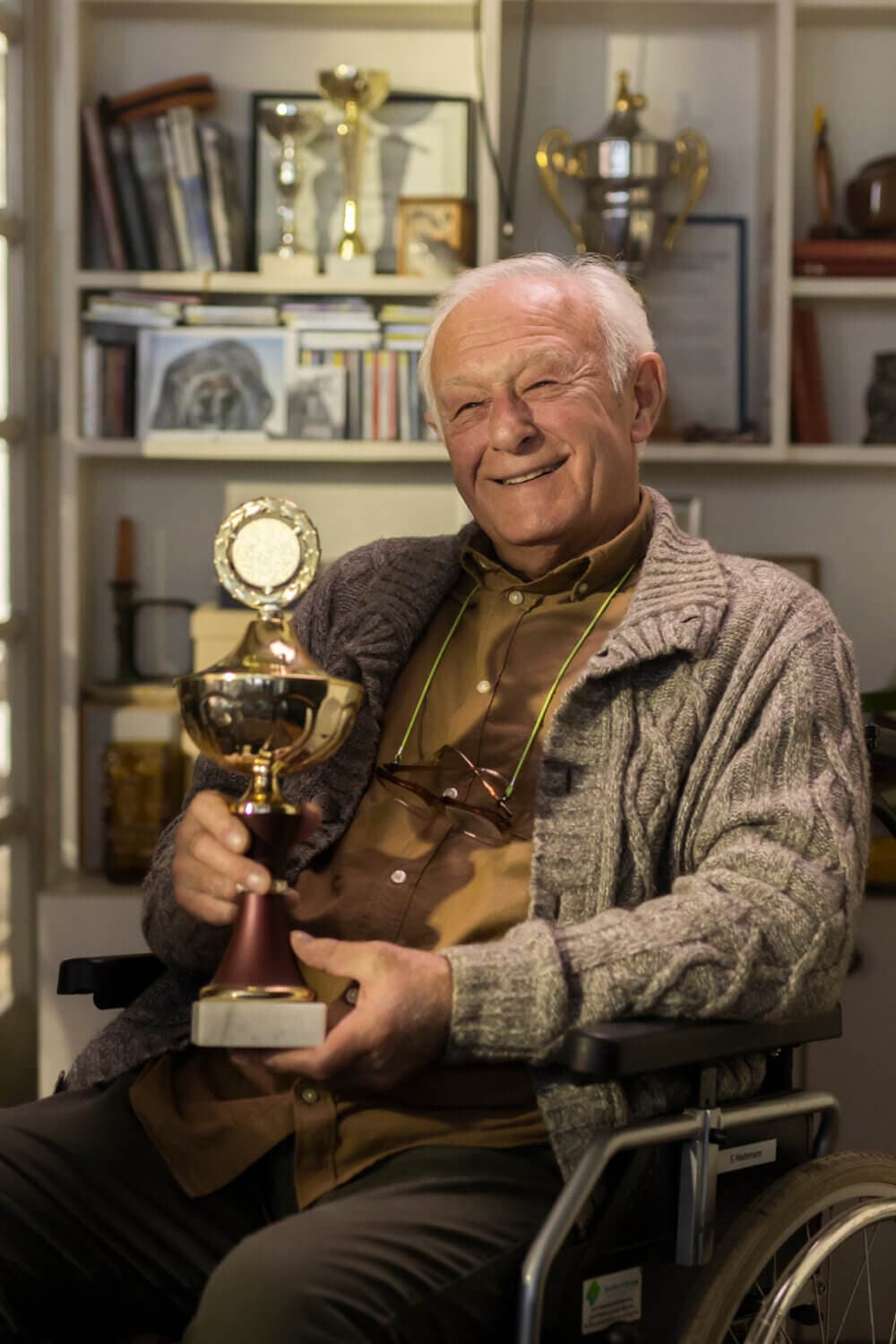 Elderly man in a wheelchair smiling and holding a trophy, with shelves of awards and books in the background. - Home Instead